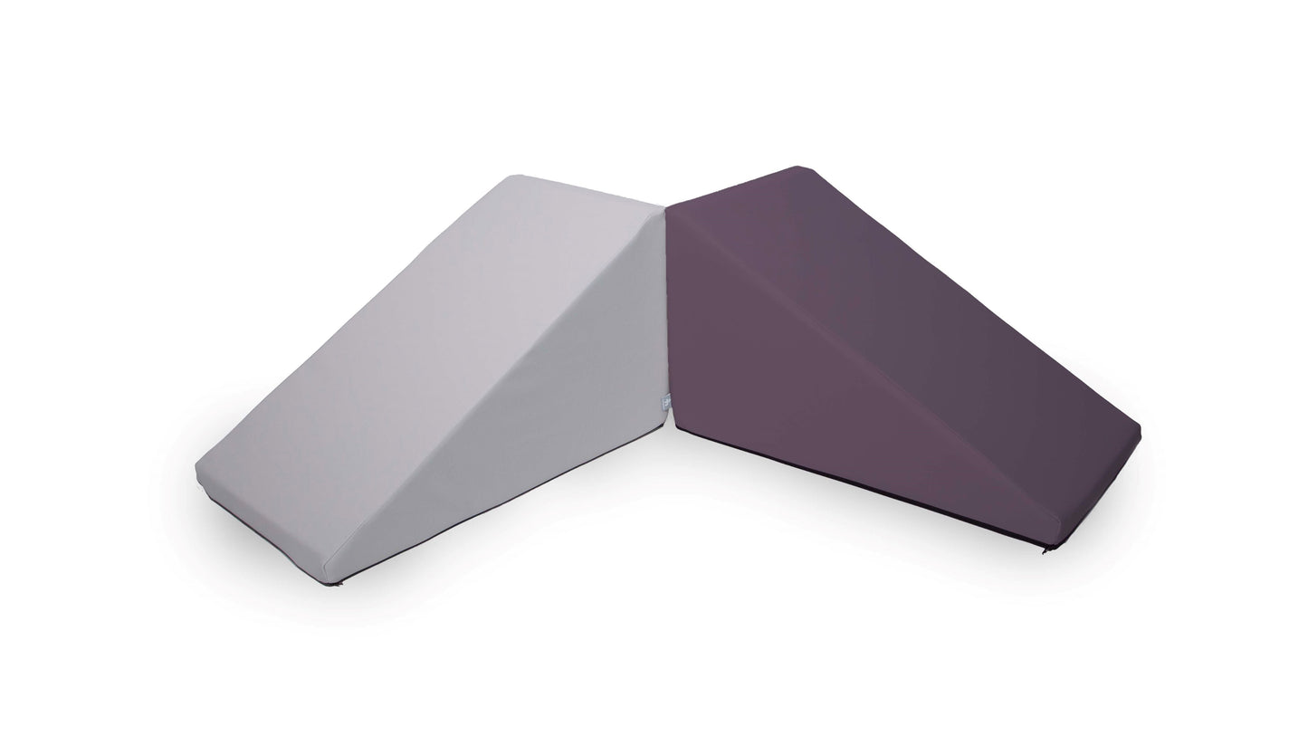 Placed side by side, two Foamnasium Mega Wedge Play Triangles, each made from CertiPUR-US certified foam, show their unique coverings: the left wedge is wrapped in light gray fabric while the right wedge boasts dark purple fabric. Both wedges feature rectangular ends and gradually slope down to a lower rectangular end.