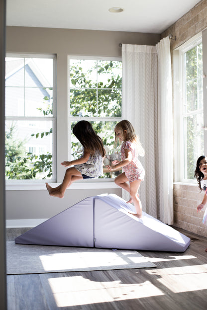 Three children are playing indoors. Two are jumping off the Foamnasium Mega Wedge Play Triangles, a durable foam playset shaped like a soft, triangular structure in the middle of a bright room with large windows. The third child watches from nearby, smiling. The room is filled with natural light, and trees are visible outside.