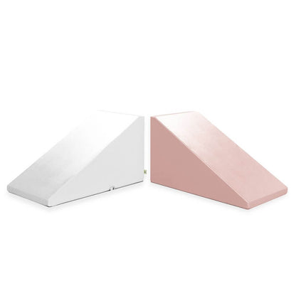 The image shows two Mega Wedge Play Triangles from Foamnasium placed side by side. The wedge on the left is white, and the one on the right is pink. Both wedges have a smooth surface and are designed to provide angled support, crafted from CertiPUR-US certified materials to ensure safety and quality.