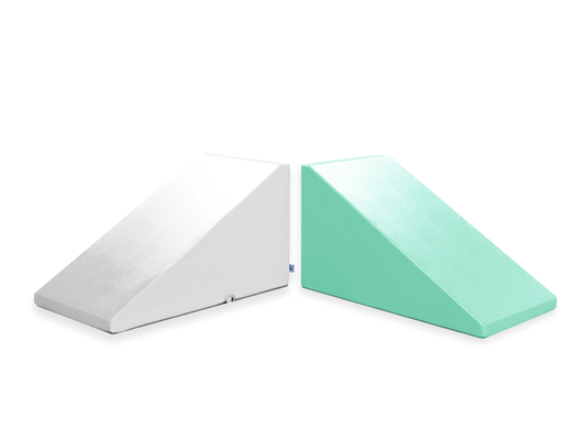 Two wedge-shaped foam incline cushions, named "Mega Wedge Play Triangles" by Foamnasium, are shown side by side. The cushion on the left is white, while the one on the right is light green. Both feature a smooth surface, are CertiPUR-US certified, and are designed for comfort and support.