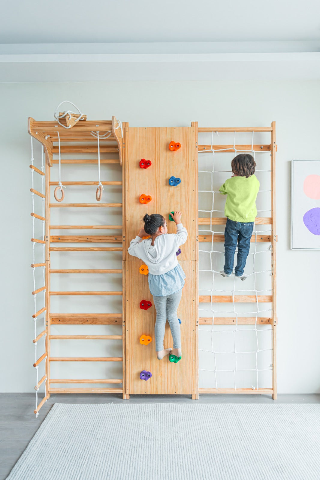 Two children are climbing the Walnut - 9-in-1 Swedish Ladder Wall Gym and Climber by PP AVENLUR in a white room with a gray carpet. The setup features climbing holds, ropes, and gymnastics rings. One child faces forward on the Swedish Ladder Wall Gym climbing holds wall, while the other ascends a rope net.